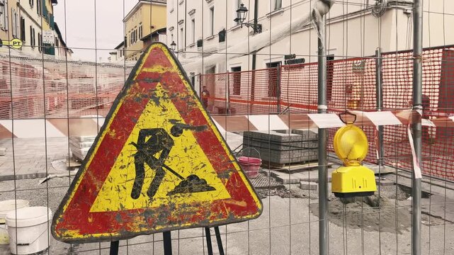 Weathered men at work warning sign standing behind a safety fence at a city street construction site with a yellow flashing beacon, indicating road closure and paving works ahead