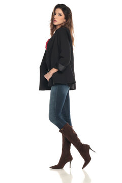 Side view of a woman in black blazer, red bra, denim jeans and high boots posing confidently, full body, studio shot on a white background