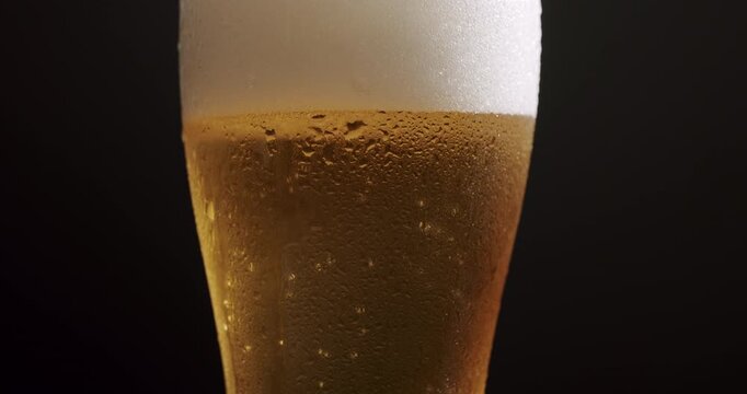 Close-up of a refreshing glass of cold light beer with rising bubbles and thick foam head, featuring condensation droplets on the exterior surface against a dark black background