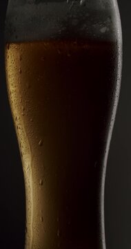 Chilled pint glass of beer slowly rotating against a dark background, showing golden color, frothy head, and condensation drops glistening under the light, creating a refreshing atmosphere. Vertical