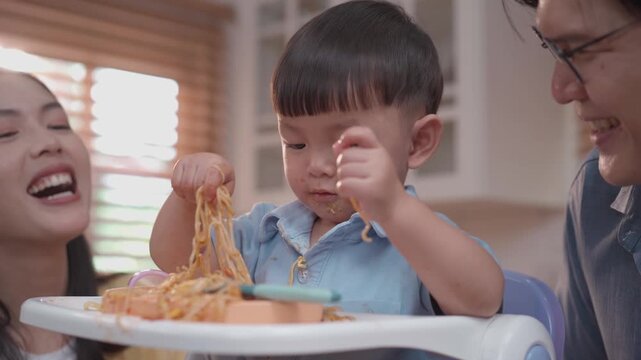 Cute toddler playing with spaghetti during mealtime, happy parents encouraging self feeding, messy eating learning, family lifestyle and child development concept
