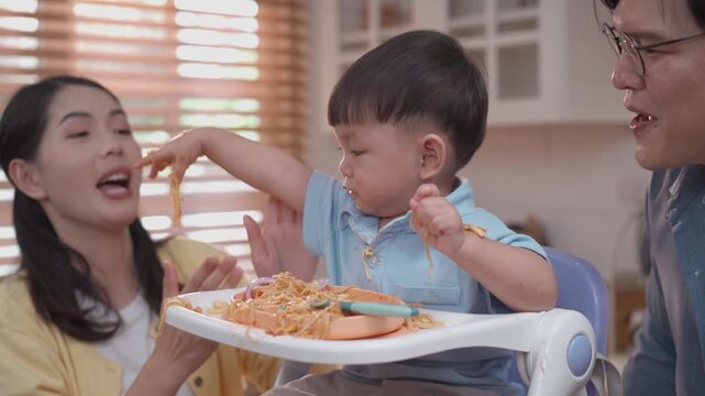 Cute toddler playing with spaghetti during mealtime, happy parents encouraging self feeding, messy eating learning, family lifestyle and child development concept