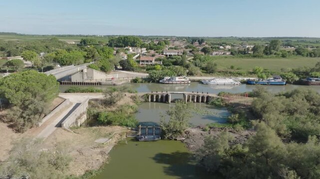 Aerial view of Camargue canal lock system with moored boats concrete structures bridge and surrounding green fields under bright blue sky