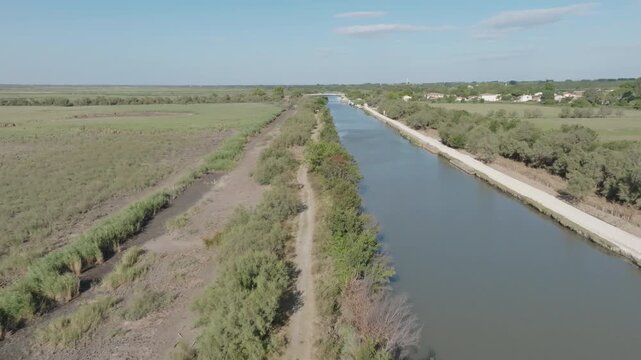 Aerial view of Camargue with long straight canal bordered by dirt path and green vegetation under bright blue sky