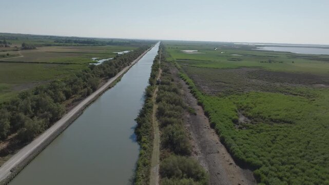 Aerial view of Camargue with long straight canal bordered by dirt path trees and green fields under bright blue sky