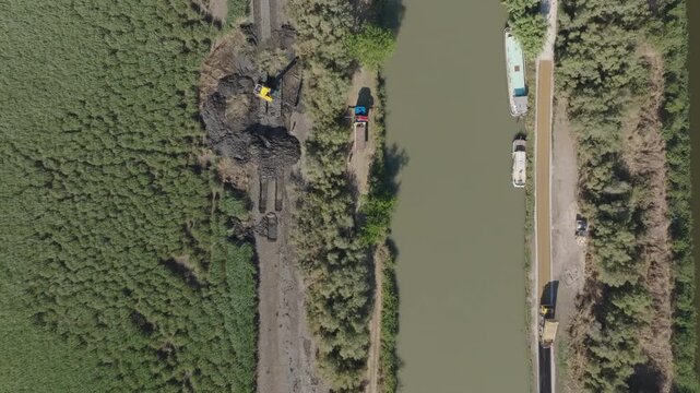 Top down aerial view of Camargue canal with yellow excavator on muddy bank and moored boats along the waterway bordered by green vegetation