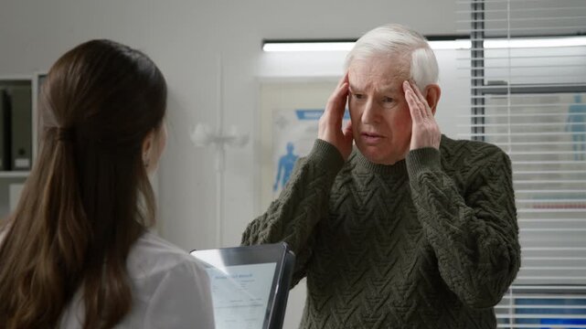 Medium shot of elderly man with silver hair describing painful headache while consulting female doctor during appointment in examination room