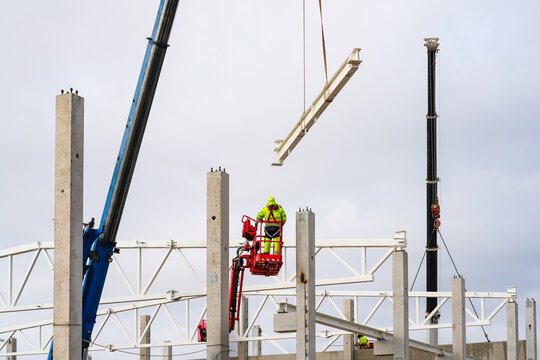 Worker in lift guides suspended steel beam as cranes position structure on concrete columns