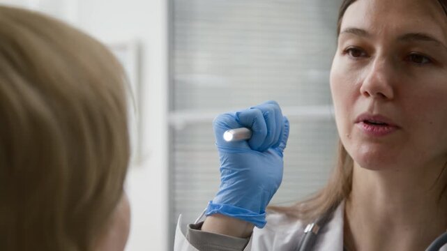 Close up shot of young adult female physician in protective gloves examining unseen patient with penlight while checking pupil reaction to light during routine check up at clinic