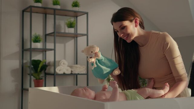 Mother entertains infant with teddy bear puppet on changing table, soft light illuminates room
