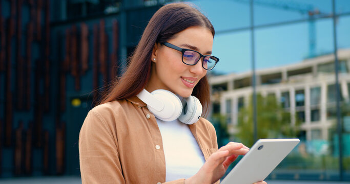 Close-up of Caucasian brunette woman in a blue shirt working on a wireless laptop outdoors in a city park, with a modern building in the background. Concept of remote work and people