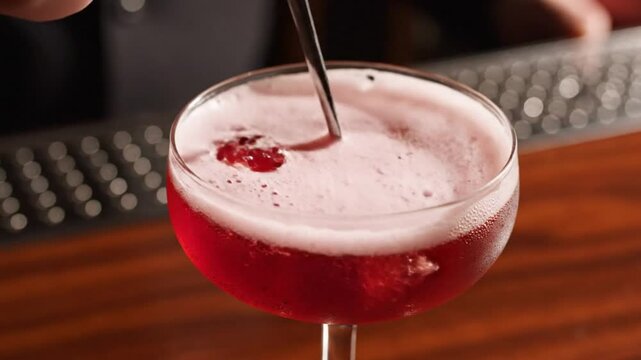 Close up of a vibrant red cocktail with a foamy top being garnished on a wooden bar top with a spoon