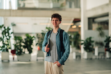 Fototapeta premium Young man standing in a bright, modern interior, looking at the camera. He is wearing a backpack and holding a tablet