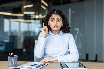 Naklejka premium Young indian businesswoman at her modern office desk has a sudden eureka moment, mouth forming an oh shape and finger raised as she discovers a solution during the workday