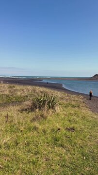 A woman with red hair stands on a black sand beach in Marokopa, Waikato, New Zealand, looking out at the ocean. The sun shines brightly on this beautiful coastal landscape.