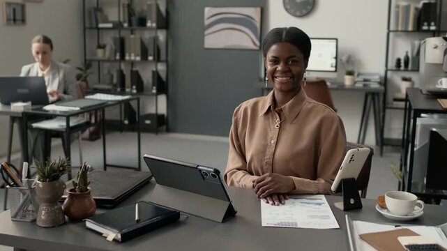 Portrait shot of young African American woman smiling for camera while sitting at office table with digital tablet and business documents