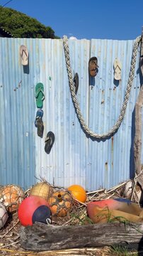 A collection of weathered fishing buoys and flip-flops are displayed against a blue corrugated metal fence on a sunny day in Marokopa, Waikato, New Zealand.