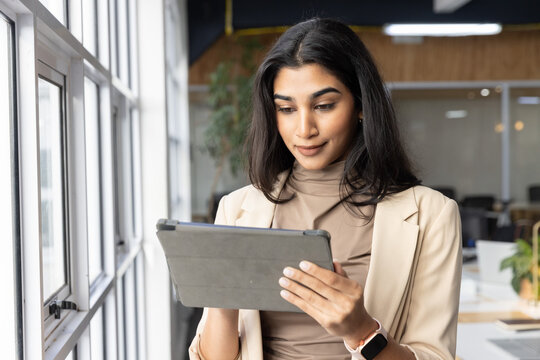 Indian woman in blazer holding tablet folio and checking screen near windows in office with watch