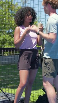 Vertical video: Walking man with paddle, ball urging woman in top sipping bottle to play on court