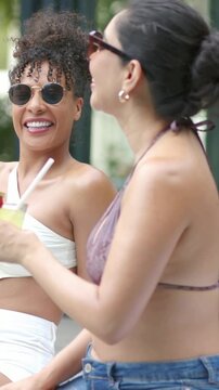 Vertical video: Sipping woman in bandeau, friend leaning offering drink with strawberry rim at pool