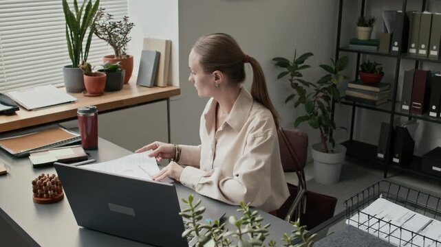 High angle shot of middle aged woman taking break from working on laptop and drinking coffee from small thermos in office