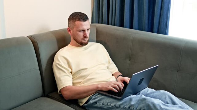 A man sits on a couch typing on a laptop. He looks focused as he works, surrounded by a simple living room setting with soft lighting.
