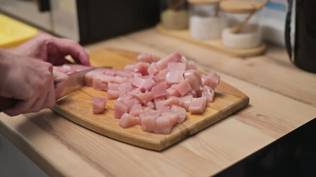 A person is chopping diced chicken on a wooden cutting board in a kitchen. The individual is focused on the preparation step of cooking, surrounded by kitchen items