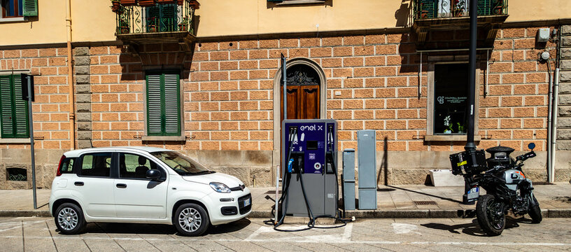 A white Fiat Panda parked next to an Enel X electric vehicle charging station on Corso Umberto I in Olbia, Sardinia, Italy, with a motorcycle nearby.
