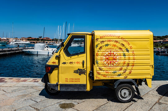 A yellow Piaggio Ape three-wheeler for Ristorante Gianni al Molo parked at the Porto Rotondo marina in Sardinia, Italy, with yachts in the background.