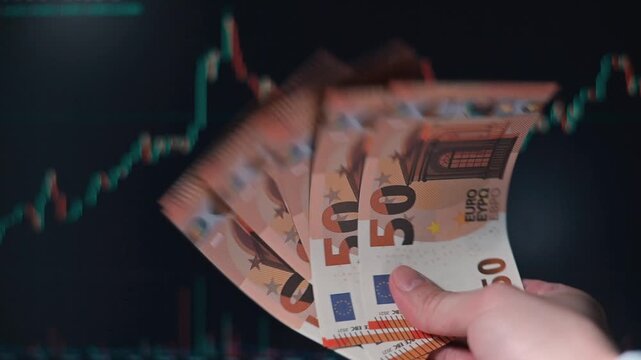 A person of unspecified gender is handling a stack of euro banknotes while analyzing stock market graphs in the background
