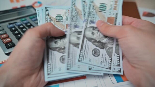 A person counts cash while seated at a desk. The workspace has financial charts, a calculator, and stacks of money visible