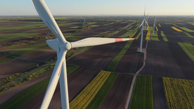Cinematic drone close-up of a large wind turbine rotor in motion, capturing the mechanical detail and height against a vast agricultural landscape at sunset.