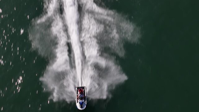 Aerial view of a jet ski with two people making a sharp turn and creating a white foamy wake on the dark green water.