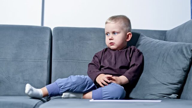 A young child sits alone on a couch and looks thoughtfully at a piece of paper. Sunlight comes in through a window, creating a warm atmosphere in the room.
