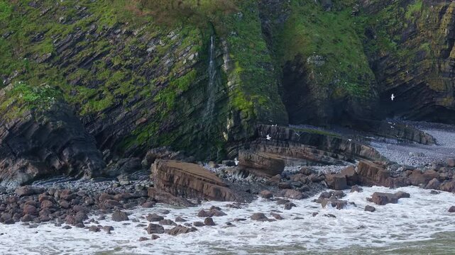 Aerial view of Mouthmill Beach which is a secluded pebble cove near Clovelly, North Devon, known for the dramatic Blackchurch Rock sea arch and waterfall. Ambient exploration by drone. 