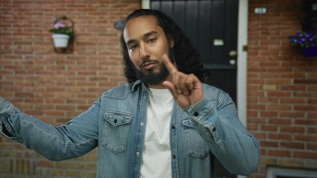 Man pointing index finger toward camera at a building entrance outside a house, wearing denim jacket and white t shirt, beard visible; assertive warning.