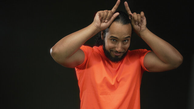 Man with fingers forming horns and bare forearms in studio with black backdrop wearing orange shirt; playful mischief.