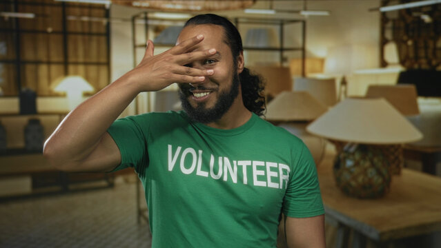 Man covering his eyes with his hand while smiling in a lamp shop building, wearing a green volunteer shirt; playful charity.