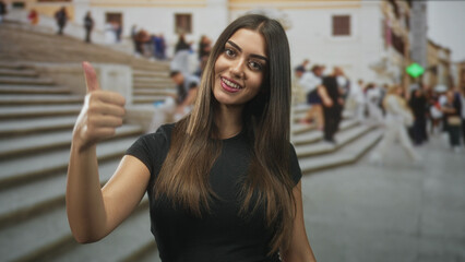 Obraz premium Smiling young hispanic woman giving thumbs up with both hands on crowded stone steps street; approval tourist moment.