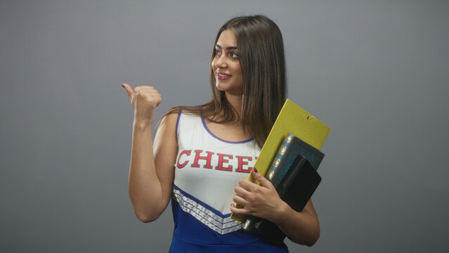 Cheerleader woman holding a yellow folder and book, thumb pointing back gesture while smiling and standing in studio; confidence.
