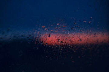 Naklejka premium Rain on glass during sunset creates a moody blue and red scene, with droplets in the foreground and a distant horizon under twilight skies as night approaches