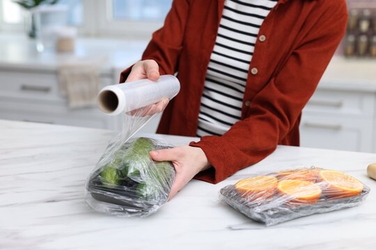 Woman wrapping container with fresh cucumbers in cling film at light marble table, closeup