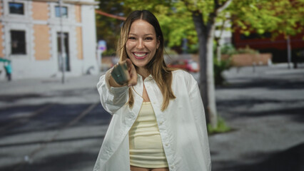 Fototapeta premium Young woman pointing finger at camera and smiling on street with building and trees in frame; playful invitation.