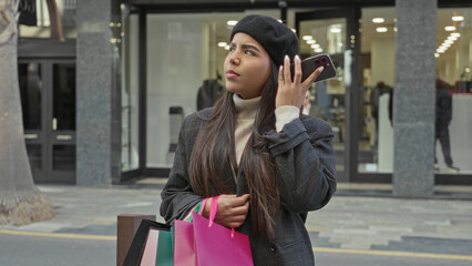 Obraz premium Young latin woman holding smartphone to ear with hand and clutching pink shopping bags outside a retail store on a busy street; frustration waiting.