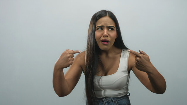Hispanic woman pointing finger to her chest and shoulder in studio wearing white sleeveless top and jeans, puzzled facial expression and accusatory pose; self doubt confusion.