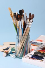 Fototapeta premium Paintbrushes in glass jar, paints and palette on light blue background, closeup