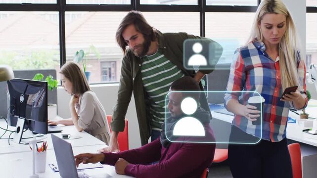 Bearded man leaning over laptop while plaid woman walking triggers UI icons over team for business