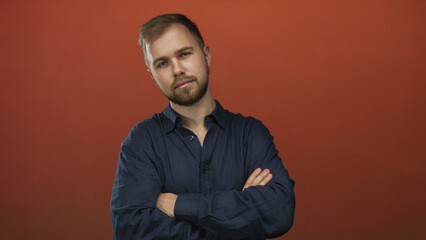 Obraz premium Young caucasian man with arms crossed in a studio against a burnt orange backdrop wearing a navy button shirt and light beard; confidence.