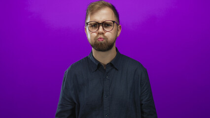 Obraz premium Man with glasses puffing cheeks in studio against a purple backdrop, young bearded and wearing dark shirt; playful moment.