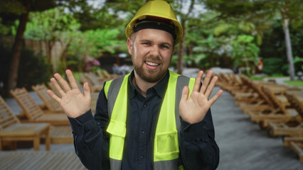 Man builder in yellow hardhat and high visibility vest stands with hands raised palms out showing open palms and an uneasy grin in building  surprise caution. © Krakenimages.com